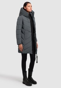 Grå pufferjakke med et chevron-design, en stor hætte og lynlås lukning. Båret med sorte leggings og ankelstøvler, der har et logo-tag.