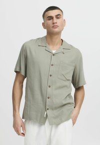 Camisa de manga corta y cuello en tela verde claro con un bolsillo y botones marrones, que presenta una textura suave y un corte relajado.