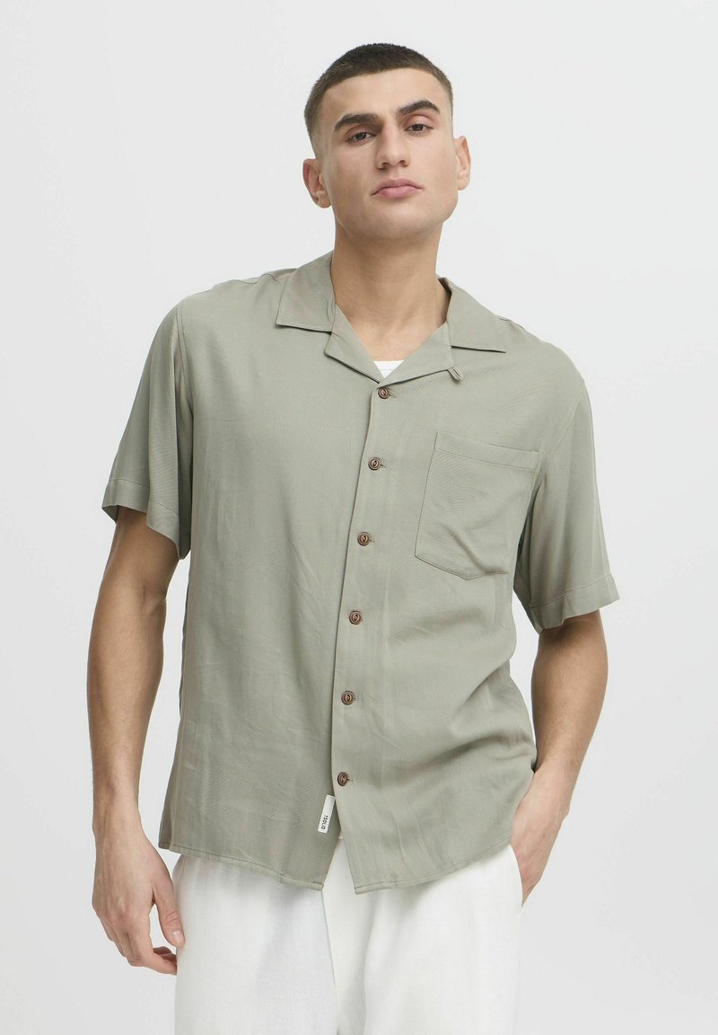 Camisa de manga corta y cuello en tela verde claro con un bolsillo y botones marrones, que presenta una textura suave y un corte relajado.