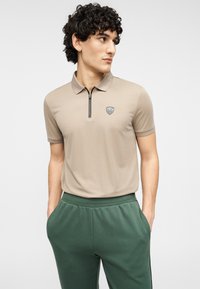 Ανδρικό κοντομάνικο πόλο σε μπεζ χρώμα με φερμουάρ στο γιακά και λογότυπο, συνδυασμένο με πράσινο παντελόνι jogger με ελαστική μέση. Μαλακή, λεία υφή υφάσματος.