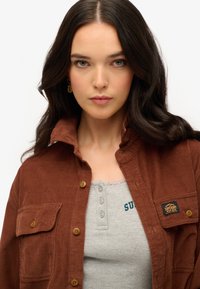 Bruine corduroy shirt met twee borstzakken en knoopsluiting, gelaagd over een grijze geribbelde tanktop met kantafwerking en geborduurde tekst.
