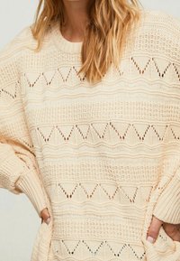 Pull en maille couleur crème avec un motif en zigzag texturé, des poignets côtelés et une coupe ample, mettant en valeur les détails de tissage ouvert.