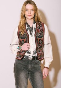 Gilet brodé avec des motifs géométriques en rouge, rose et or sur un fond noir, porté sur une blouse blanche et un jean foncé.