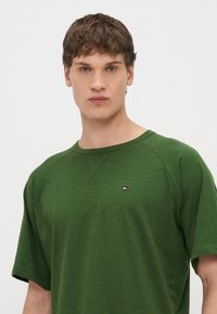 Σκούρο πράσινο κοντομάνικο T-shirt με στρογγυλή λαιμόκοψη και ραγκλάν μανίκια. Φέρει μικρό λογότυπο σε κόκκινο, άσπρο και μπλε στο στήθος.