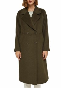 Kobieta ubrana w długi oliwkowozielony wełniany płaszcz dwurzedowy z szerokimi klapami, nałożony na pasek w paski i białe spodnie, na białym tle.