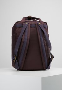 Ciemnobrązowy plecak z regulowanymi, wyściełanymi szelkami i uchwytem u góry, prezentowany na białym piedestale na szarym tle.