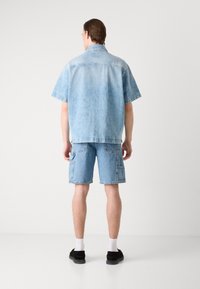 Camisa de mezclilla azul clara y pantalones cortos, corte relajado con mangas cortas, costuras visibles, bolsillos parches en los pantalones cortos, llevados con calcetines blancos y zapatos negros.