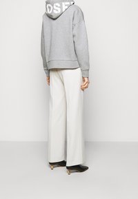 Persoon in een grijze hoodie met witte tekst op de capuchon, crèmekleurige wijde broeken en zwarte hoge hakken, staand tegen een plain witte achtergrond.