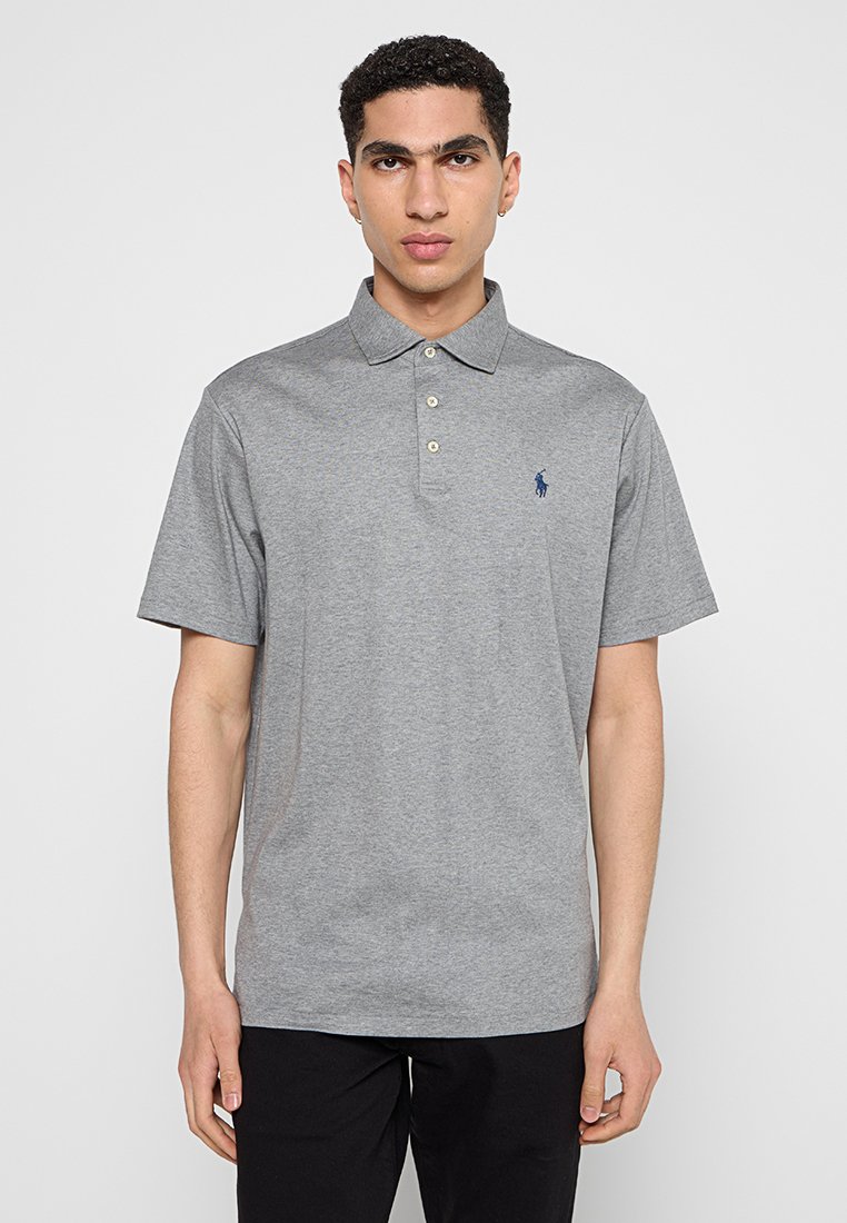 Joven vestido con una camiseta polo de manga corta gris con un pequeño logo azul marino en el lado izquierdo del pecho, de pie frente a un fondo liso.
