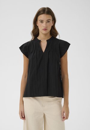 Donna che indossa una blusa nera senza maniche con scollo a spacco e pantaloni crema a vita alta, in piedi davanti a uno sfondo semplice.