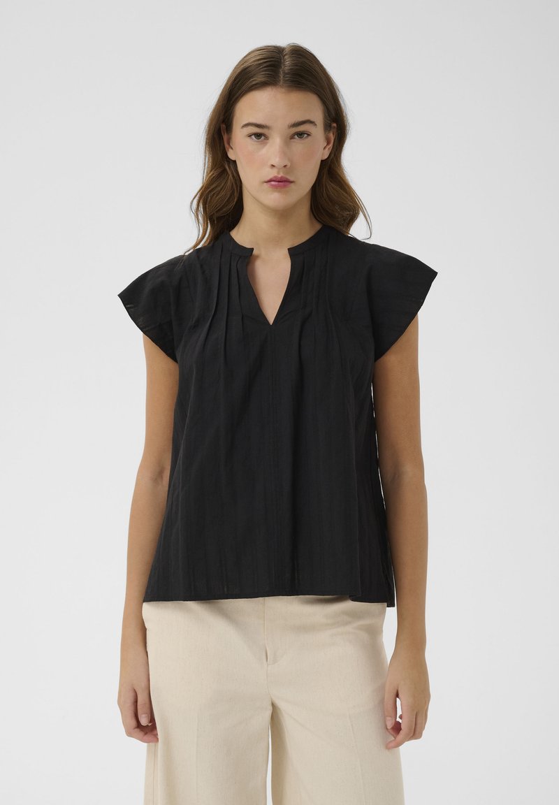 Femme portant un chemisier noir sans manches avec une encolure fendue et un pantalon crème taille haute, debout devant un fond uni.