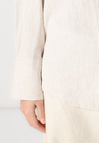 Gai bēša lina krekls ar nedaudz raksturīgu virsmu, ar pielāgotu aproci un brīvu piegriezumu, apvienots ar krēmveida kokvilnas biksēm.