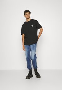 Schwarzes Baumwoll-T-Shirt mit rundem Halsausschnitt, mit einem weißen Logo auf der linken Brust. Kombiniert mit hellblauen Jeans und schwarzen Stiefeln.