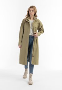 Lange olijfgroene jas van lichtgewicht stof, met een hoge kraag, drukknoopsluiting en zijzakken met ritssluiting, gedragen over lichte jeans en laarzen.