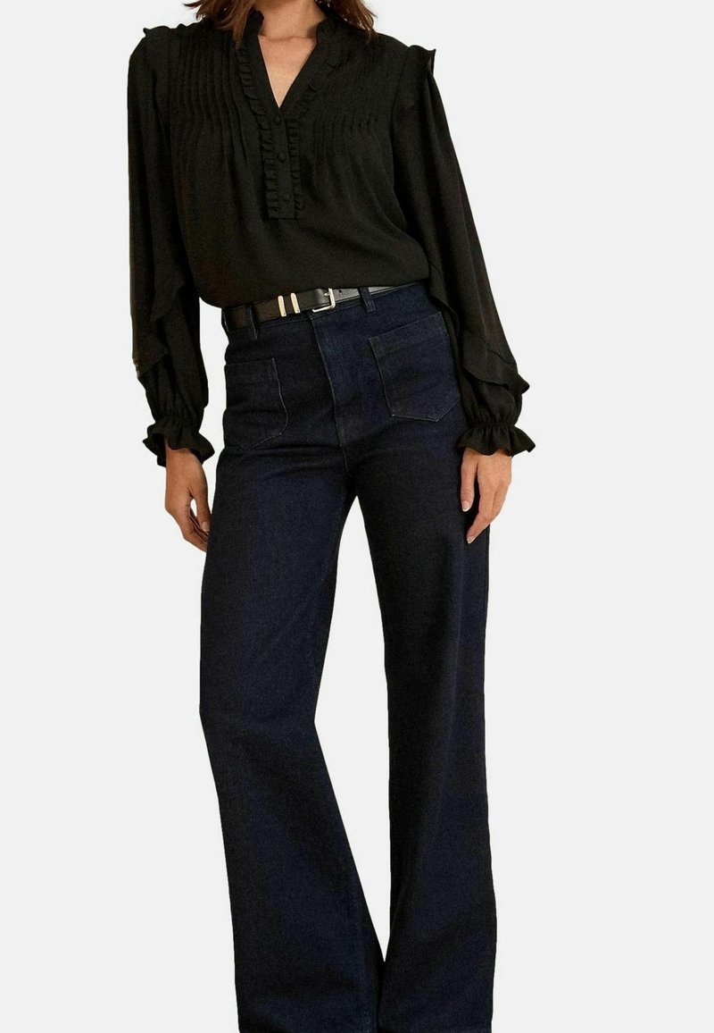Femme vêtue d'un chemisier noir à manches longues avec des volants et un pantalon large bleu foncé taille haute avec une ceinture noire.
