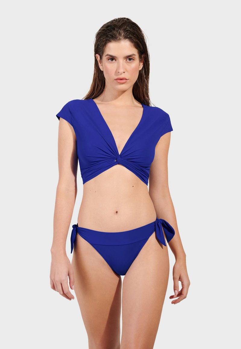 Plavi bikini set koji se sastoji od krojene majice s mašnom i kapicama te odgovarajućih donjih gaćica s vezanjem sa strane. Glatka, rastezljiva tkanina jednobojne boje.