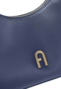 Granatowa skórzana torebka o zaokrąglonym kształcie, z złotym zamkiem błyskawicznym i wygrawerowanym złotym logo na froncie. Gładka tekstura.