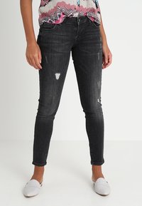 Jeans skinny negros de denim con detalles desgastados, que presentan un ajuste slim y una cintura de altura media. Llevados con una blusa de patrón colorido y zapatos blancos.