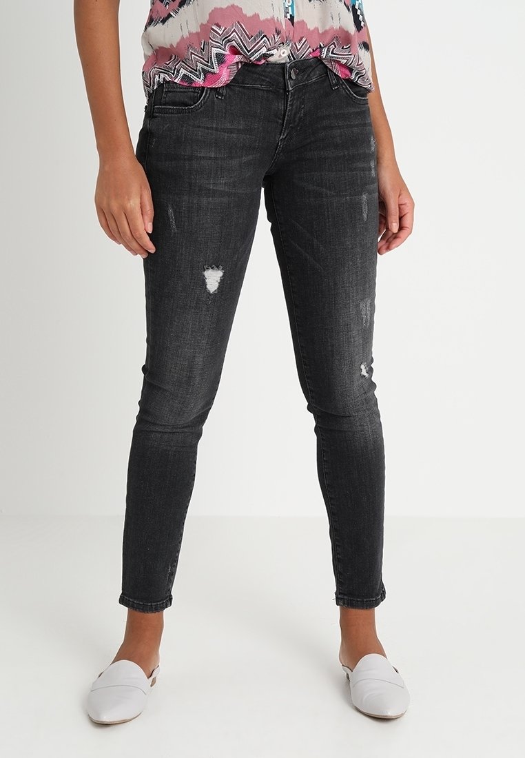 Jeans skinny negros de denim con detalles desgastados, que presentan un ajuste slim y una cintura de altura media. Llevados con una blusa de patrón colorido y zapatos blancos.