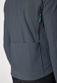 Chaqueta de ciclismo gris con una textura suave, que cuenta con un bolsillo trasero y un bolsillo lateral con cremallera. Nombre de la marca "Isadore" impreso verticalmente.