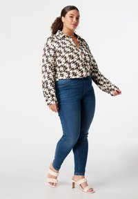 Blouse à col avec un motif géométrique en noir et beige, associée à un jean skinny bleu foncé et des escarpins à brides tressées claires.