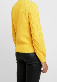Pull tricot jaune avec motif torsadé texturé sur les manches et bord côtelé. Associé à un pantalon en simili cuir noir pour un contraste.