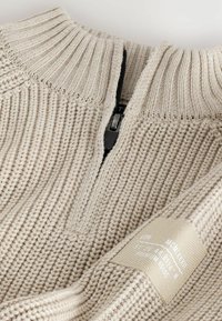 Beige gestrickter Pullover mit einem halben Reißverschluss-Kragen, der über eine gerippte Textur und ein Etikett-Detail verfügt. Der Stoff wirkt weich und atmungsaktiv.
