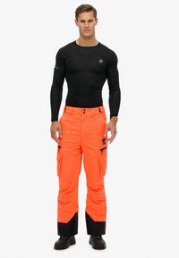 Man staat in een aansluitend zwart langemouwenshirt, feloranje cargobroek met zwarte vlekken, en zwarte laarzen voor een witte achtergrond.