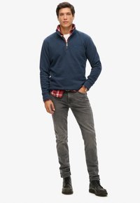 Pullover in pile con zip color navy sopra una camicia a quadri rossa, abbinato a jeans grigi e stivali neri. Presenta un piccolo logo sul petto.