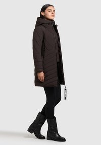 Brun quiltet parka med hætte, der har diagonale syninger, frontlynlås og sidelommer. Sorte leggings og knæhøje sorte støvler fuldender looket.