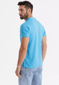 Světle modré polo tričko vyrobené z bavlny, s límečkem a krátkými rukávy. Zadní pohled ukazuje boční rozparky a jednoduchý, čistý design.