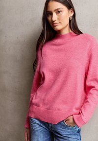 Maglione rosa lavorato a maglia con colletto alto, polsini e orlo a coste. Abbinato a jeans in denim blu con vestibilità rilassata e tasche anteriori.