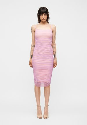 Γυναίκα φορώντας εφαρμοστό, ριχτό λιλά μίντι φόρεμα με λεπτές τιράντες, όρθια με ουδέτερη έκφραση και ορατά τατουάζ στα χέρια.