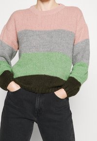 Persona che indossa un maglione spesso a maglia con ampie righe orizzontali nei toni pastello di rosa, grigio, verde e marrone scuro, insieme a jeans neri, mani nelle tasche.