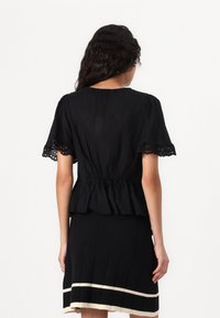 Svart peplum topp med korte, volangede ermer og utskjæringsdetaljer. Laget av lett stoff, har en samlet midje og subtil tekstur.