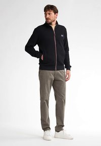 Musta vetoketjullinen takki, jossa on kuvioitu pinta, valkoiset yksityiskohdat ja sivutaskut, yhdistetty vaaleanvihreisiin chinoihin ja valkoisiin tennareihin.