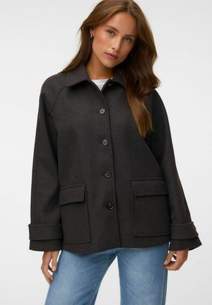 Femme aux longs cheveux bruns portant une veste gris foncé boutonnée avec de grandes poches avant et un jean bleu clair sur un fond uni.