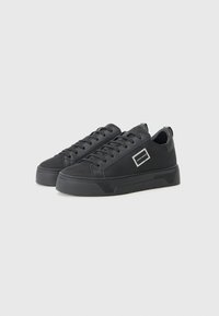 Schwarze Sneakers mit einem strukturierten Stoffobermaterial, runder Zehenpartie, flacher Sohle und Schnürdesign. Verfügt über ein rechteckiges Etikett an der Seite.
