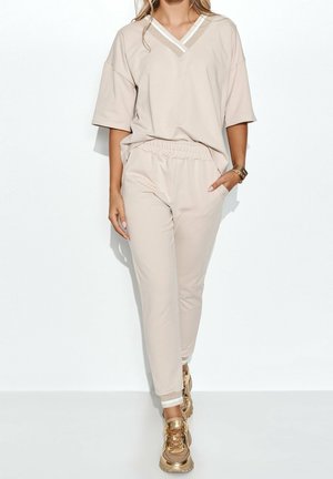 Kobieta w beżowej bluzce z krótkim rękawem i dekoltem w V oraz pasujących spodniach z elastycznym paskiem, w złotych adidasach i akcesoriach na nadgarstki, stojąca na tle białej ściany.