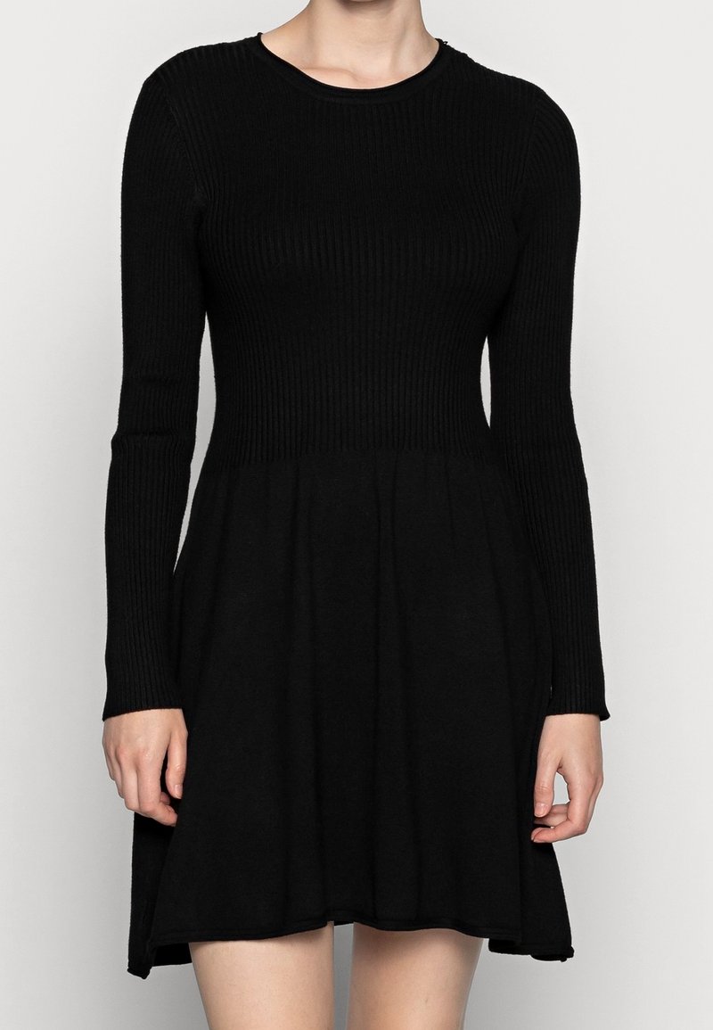 Robe noire tricotée à manches longues avec une texture côtelée, décolleté rond, corsage ajusté et jupe évasée. Tissu lisse, design classique.