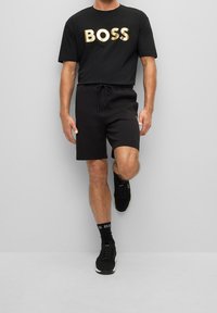 Man med svarta shorts, svart t-shirt med guldtexten "BOSS", svarta sneakers och svarta strumpor som står på ett ben mot en grå vägg.