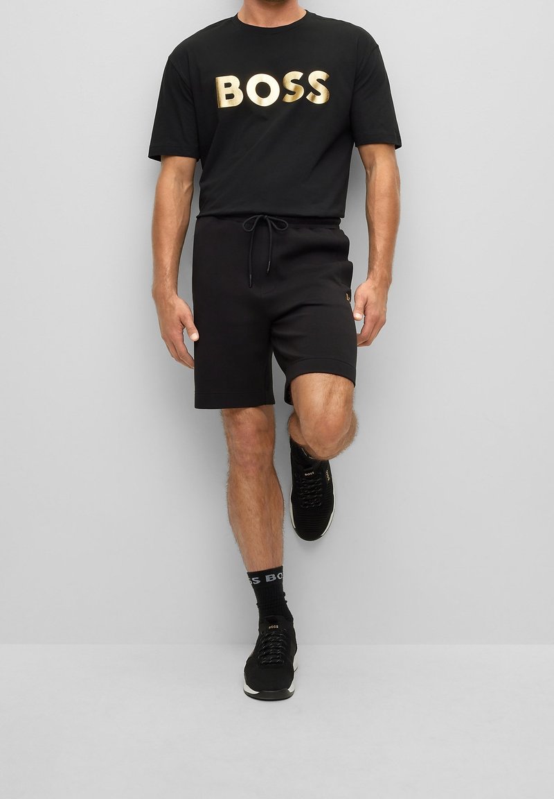 Man med svarta shorts, svart t-shirt med guldtexten "BOSS", svarta sneakers och svarta strumpor som står på ett ben mot en grå vägg.