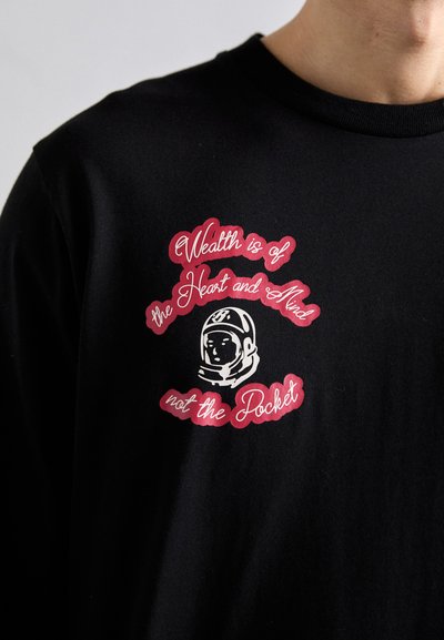 Black shirt with white astronaut helmet graphic and red text stating "Wealth is of the Heart and Mind not the Pocket" on the chest.