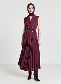 Gilet double boutonnage bordeaux avec ceinture à la taille, associé à une jupe plissée. Tissu lisse avec un léger éclat. Bottes à talons montantes.