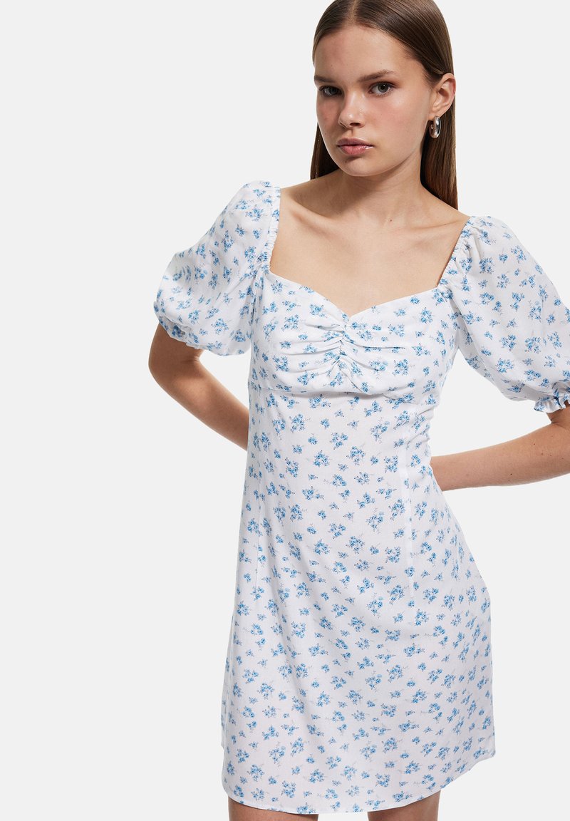 Vestido floral hecho de una tela ligera con escote cuadrado, mangas abullonadas y un fondo blanco que presenta pequeñas flores azules.