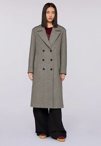 Cappotto doppiopetto in lana grigia con grandi bottoni neri, rever a incavo e design che arriva al ginocchio, abbinato a pantaloni neri ampi.