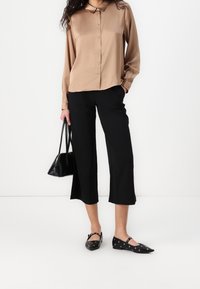 Lichtbruine satijnen shirt met lange mouwen, zwarte cropped broeken, zwarte handtas en zwarte platte schoenen met oogjes. Glad textuur en getailleerde pasvorm.