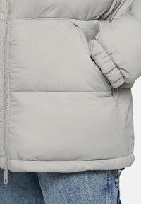 Lekka szara kurtka puchowa z suwakiem z przodu, wykonana z teksturowanego materiału, z elastycznymi mankietami i boczną kieszenią. Połączona z dżinsami.