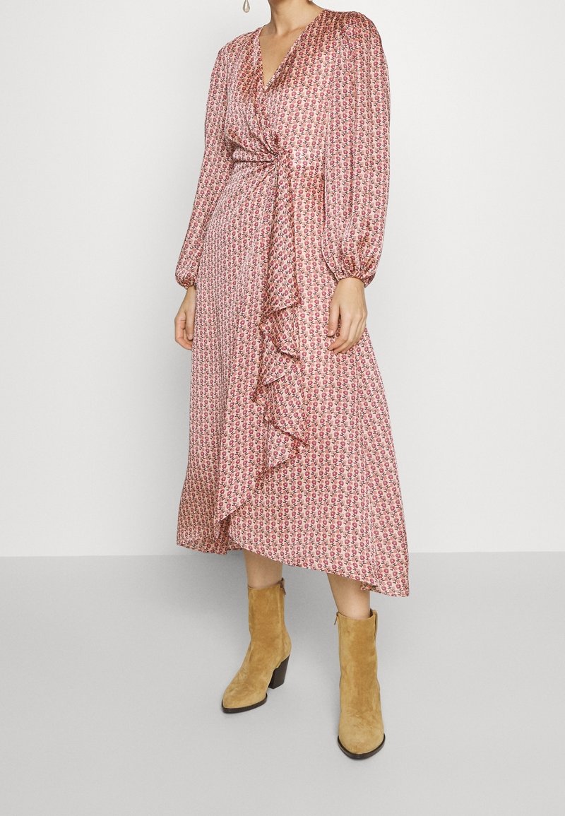 Femme portant une robe portefeuille rose à fleurs avec manches longues et détail à volants, associée à des bottines en daim beige.