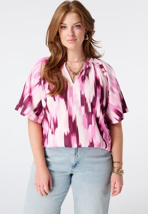 Femme aux longs cheveux roux porte un chemisier à manches courtes à motifs rose et blanc, un jean bleu clair, des bracelets en or et des colliers superposés.